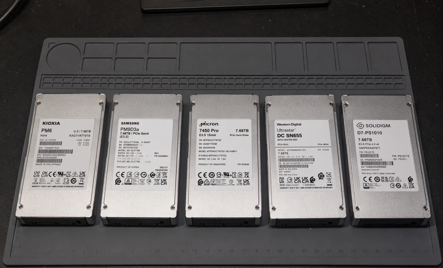 Photorealistic photograph of five real E3.S enterprise NVMe SSDs lined up in a row on an anti-static mat, dark lab bench.